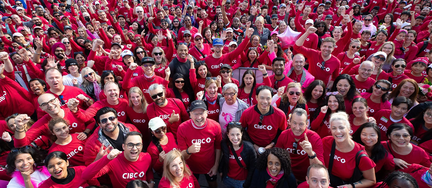 A large group of people wearing matching red CIBC shirts gather closely together, many raising their fists or arms in the air in a gesture of solidarity and enthusiasm. The crowd appears to be participating in a community or corporate event, demonstrating unity and team spirit.